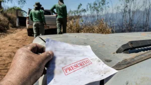 Auto de infracao ambiental segurado por mao suja de terra sobre capo de caminhonete, com carimbo vermelho PRESCRITO, pasto ao fundo