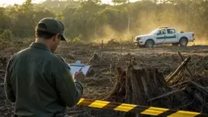 Agente de fiscalização de costas anota em prancheta diante de área desmatada, com caminhonete branca ao fundo no fim da tarde.