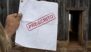 Mão suja de terra segura, na porta de um galpão rural, documento com carimbo vermelho PRESCRITO sobre a multa ambiental